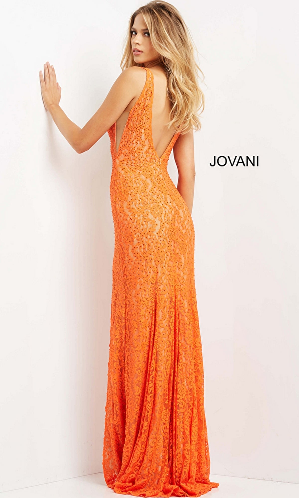 A woman faces a wall in the Long Prom Dress 08674 by Jovani—a bright orange, sequined, sleeveless gown with a deep open back. The floor-length evening dress sparkles as